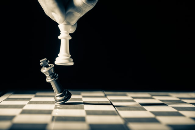 A hand moving a white chess piece to make the back king fall on a chess board.