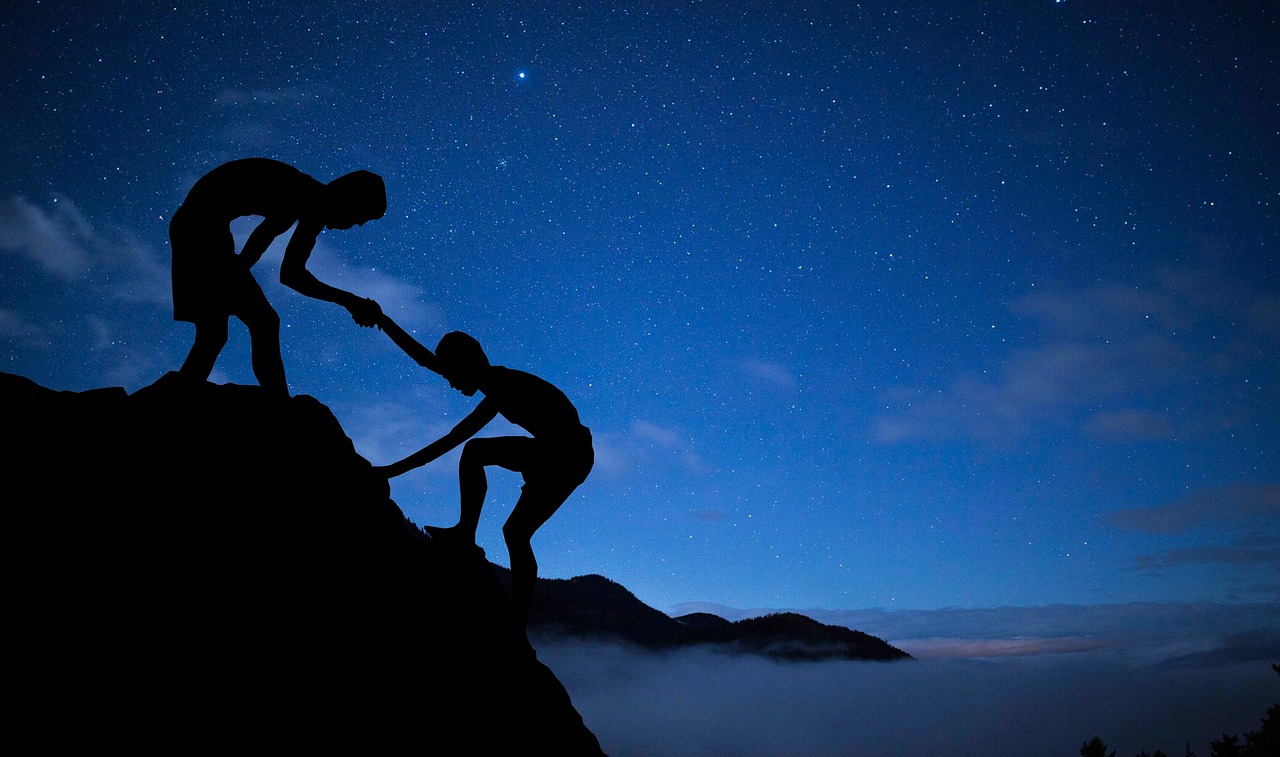 The silhoutte of a man pulling up another man while rock climbing