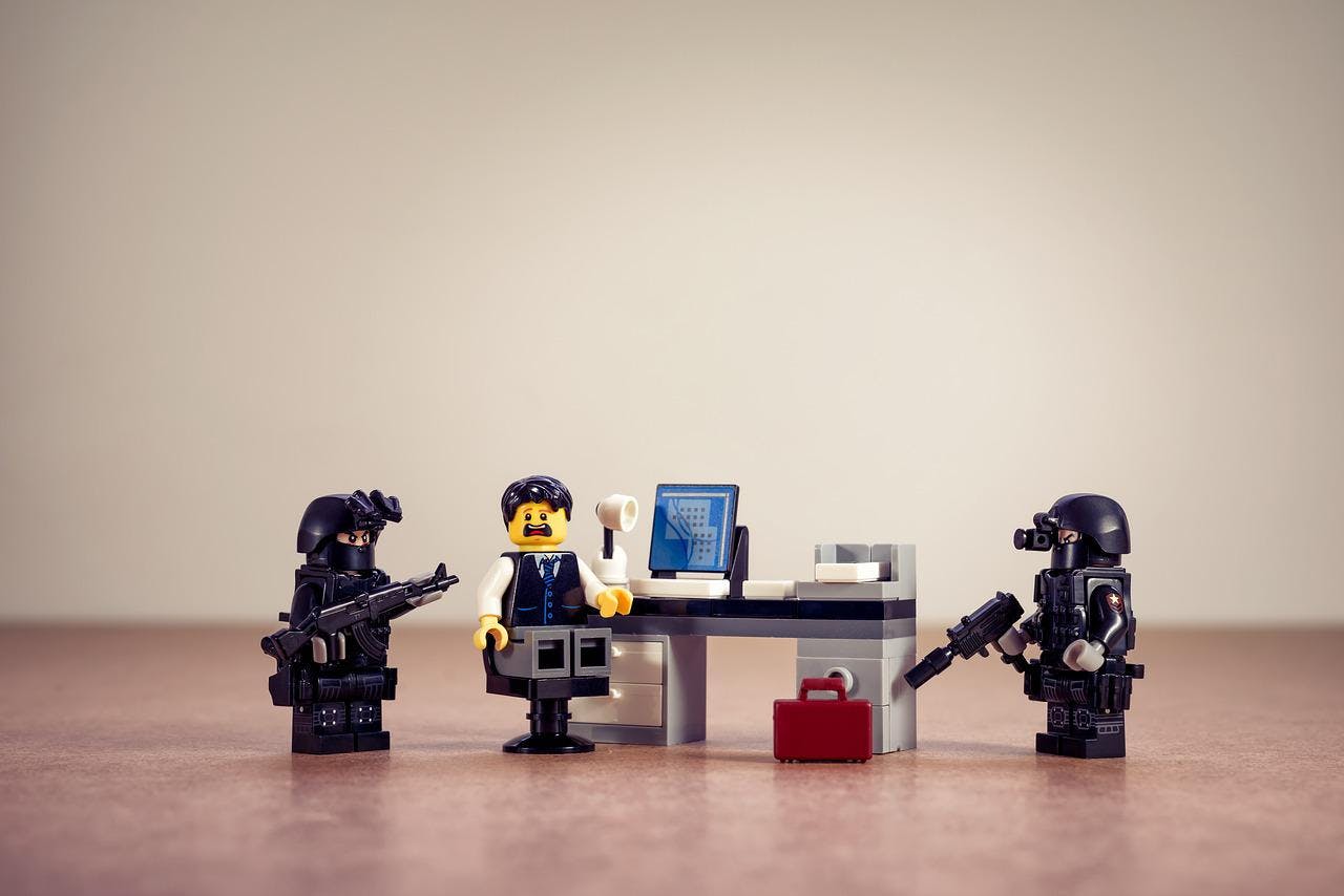 A picture of a lego piece sitting at a lego computer desk, surrounded by other gun-wielding lego pieces