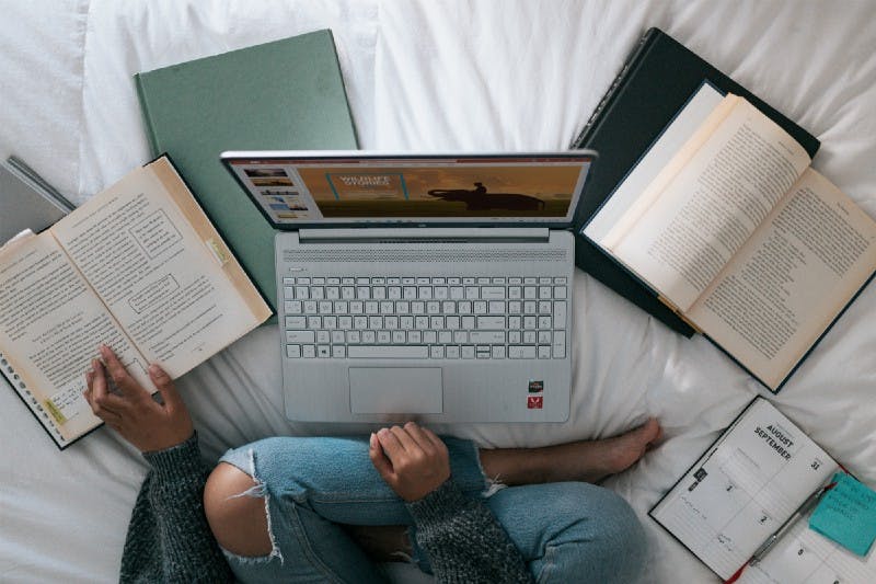A woman sitting cross-legged and barefoot on a bed while on a laptop and referencing multiple open books splayed around the bed