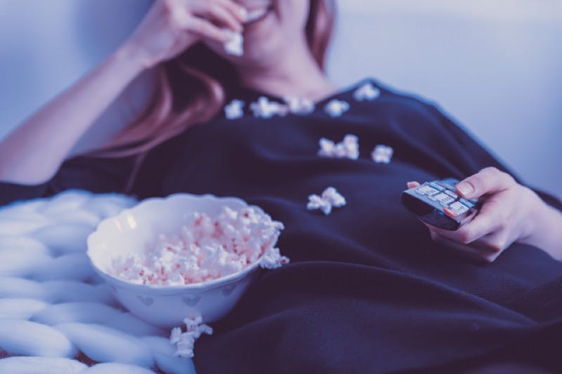A reclined woman watching TV, holding a remote control, and laughing while eating popcorn