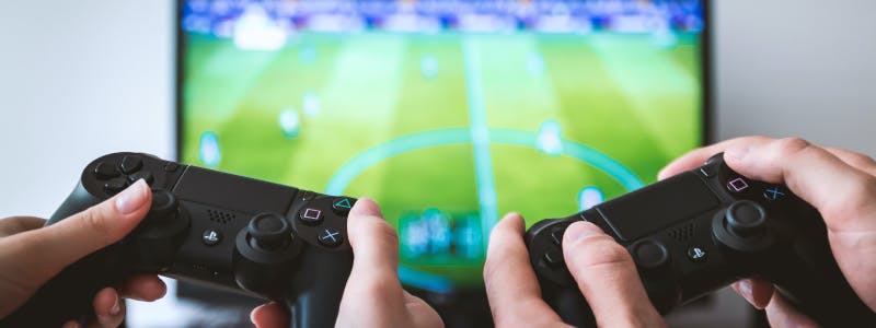 Two PS5 controllers, one held by two female hands and one held by two male hands, as they play a soccer game on a television in the background, representing the variety in target audience