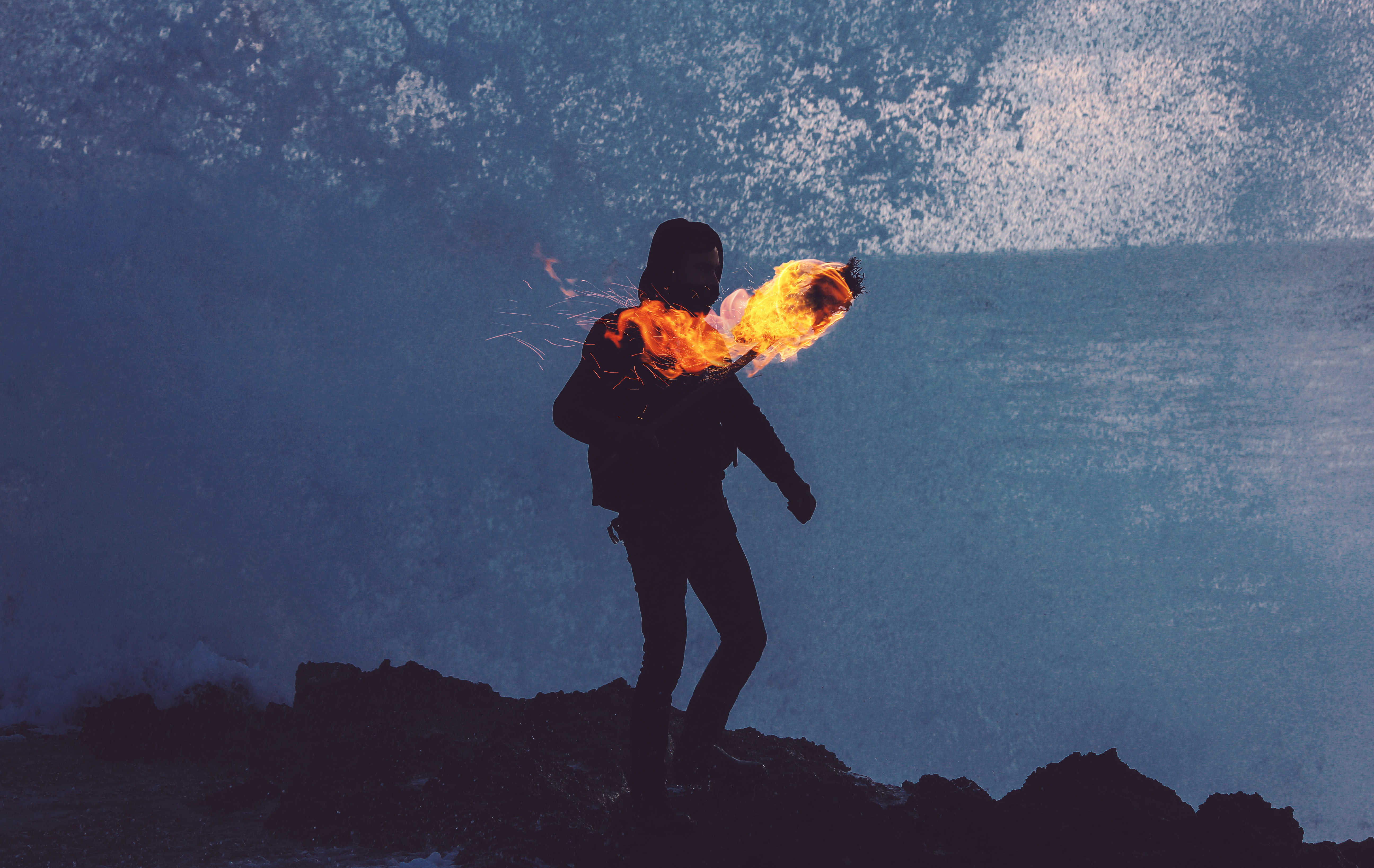 A sillouhette person with a fire torch in their hand, walking over rocky terrain. By Aziz Acharki on Unsplash