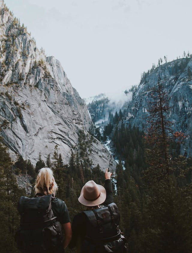 A mountain gorge with steep rock cliffs either side. In the foreground, two people are hiking, with large backpacks on. One, wearing a hat, points the way they need to go. Photo by @von_co Ivana Cajina on unsplash.com   