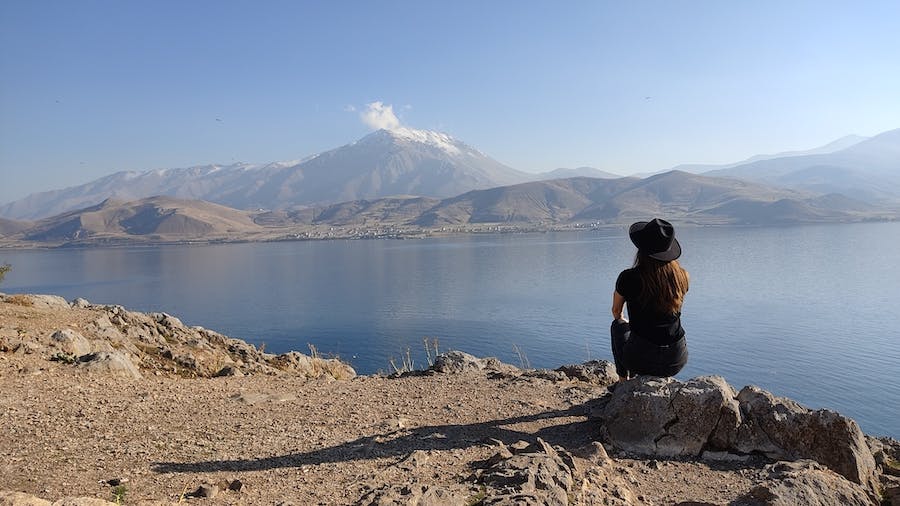 kobieta w kapeluszu siedzi na kamieniu i patrzy na jezioro i góry