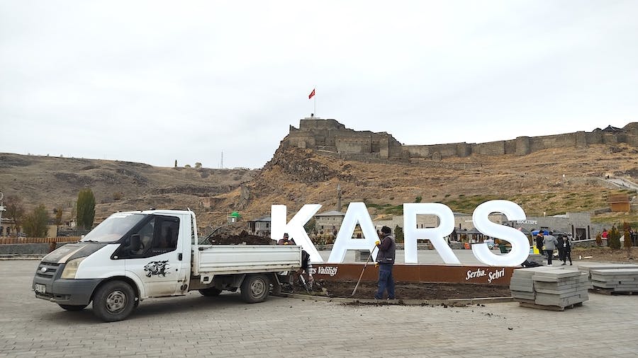 napis Kars, samochód i robotnicy naprawiający chodnik, w tle cytadela w Kars z turecką flagą 