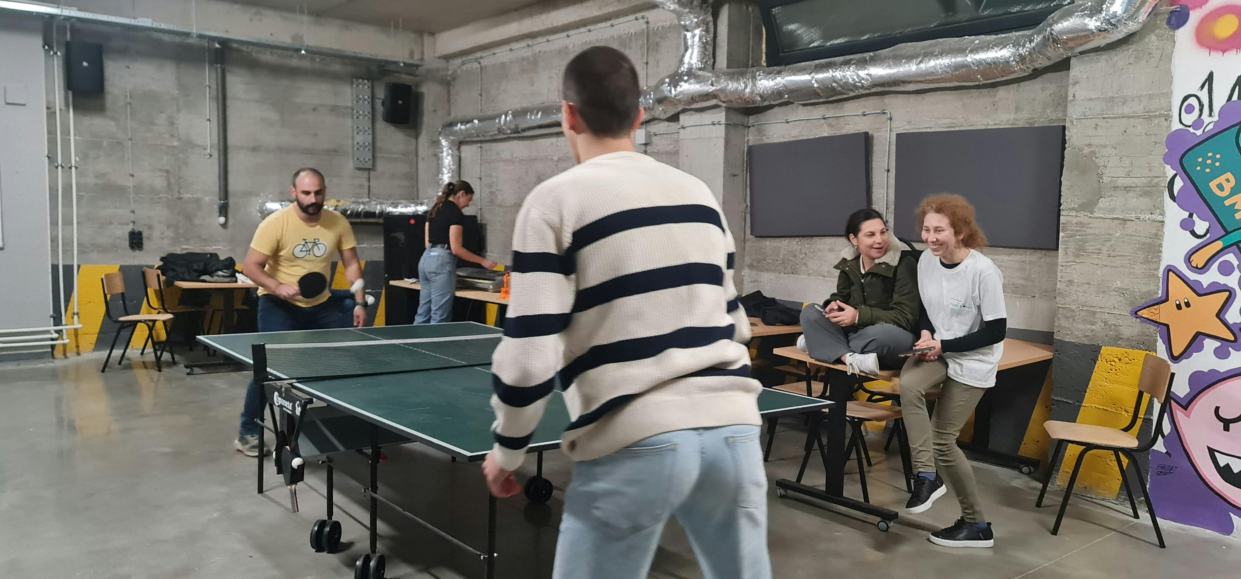 This image contains two players playing ping-pong on a table. There is one person standing behind them and two persons having a conversation. The area is filled with chairs and desks. 