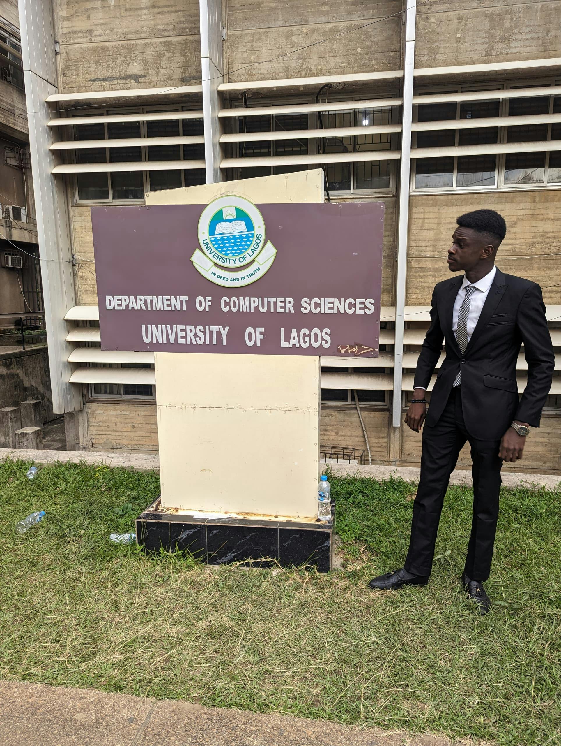 Sunkanmi dressed in a black suit with a black and silver colored tie standing next to the university of Lagos, Computer Science department sign board