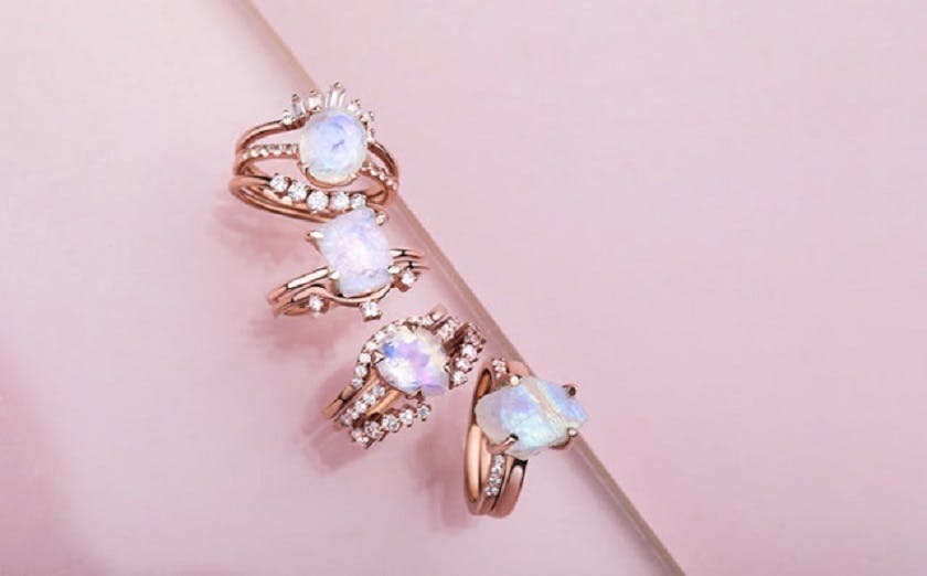 A close-up photo of a stack of five Moonstone rings with round, white gemstones on a light pink surface.