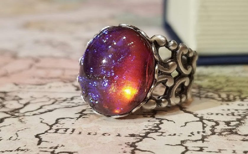 Close-up of a silver ring with a purple and orange stone, possibly a fire opal, on top of a unfolded map. Text on the image reads “Dragon’s Breath Opal Ring” and “Vintage Sterling Silver Dragon’s Breath Ring”.