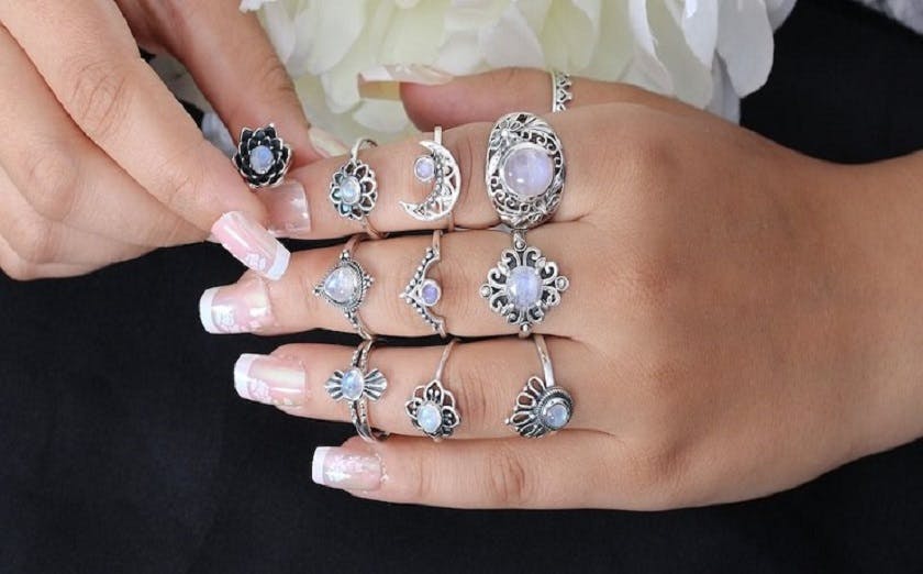 Close-up of two hands adorned with various ornate silver rings featuring Moonstone gemstones and intricate designs, alongside a subtle manicure with iridescent nail polish, partially holding a white flower.