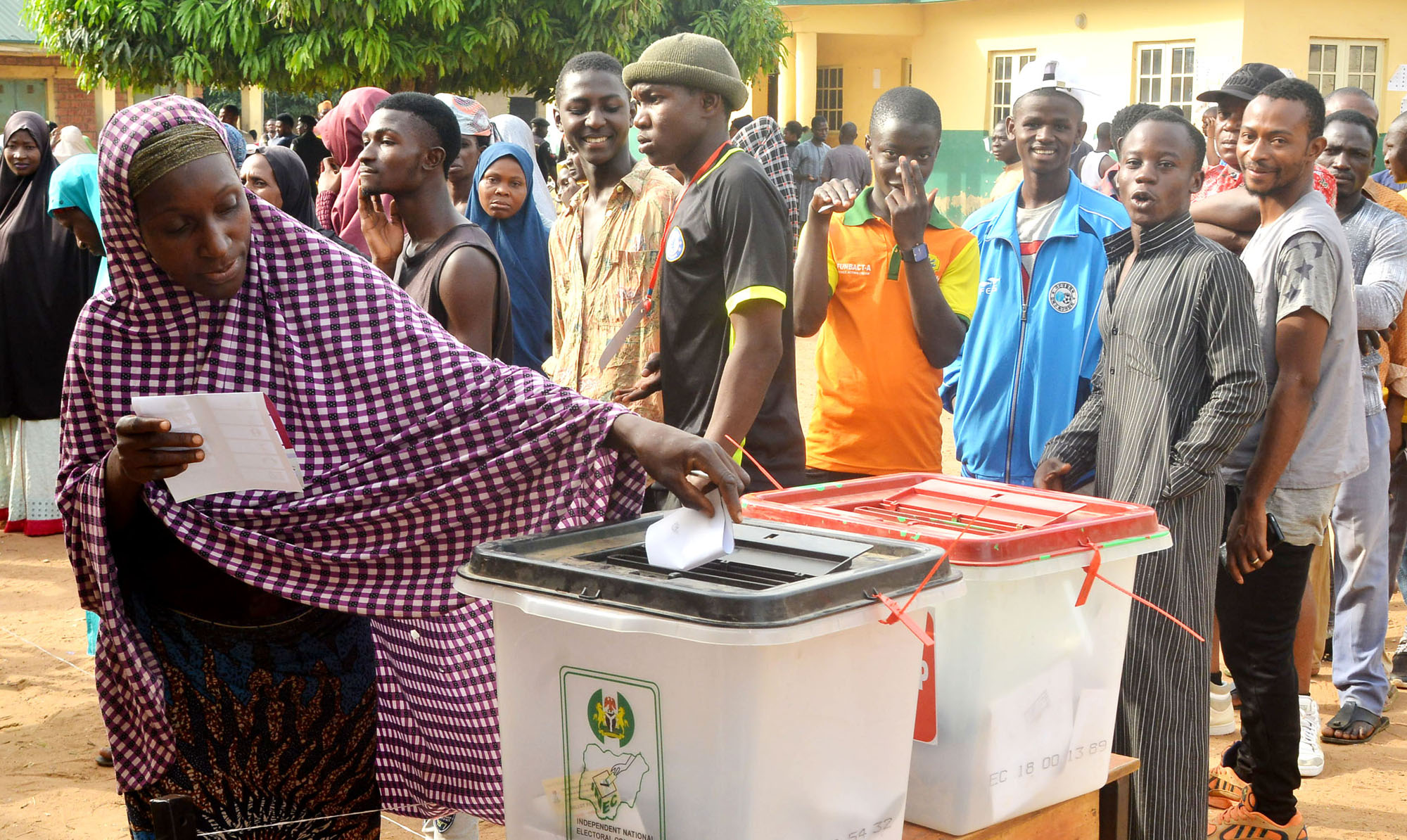 A woman casts her vote into a ballot box during an election while a group of people wait in line behind her. The scene takes place outdoors, with many people wearing colorful clothing and headscarves.