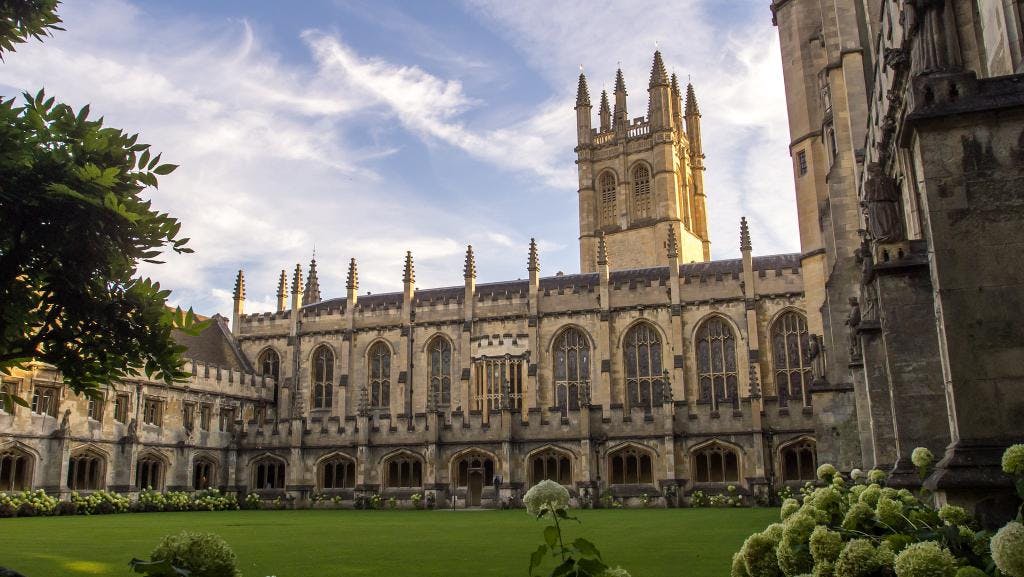 A historic stone building with tall arched windows and a tower, surrounded by a courtyard with green grass and shrubs. The sky is partly cloudy. Best place for international student.