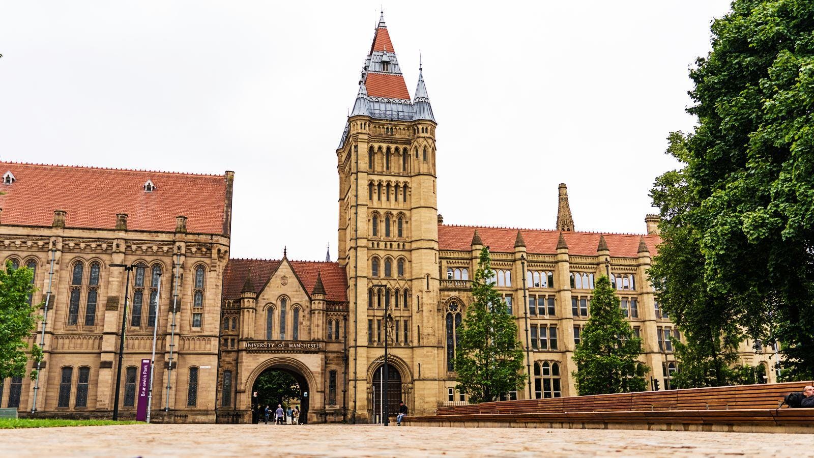 A historic building with a tall clock tower and red tiled roofs, surrounded by trees and a paved courtyard. Best place for international student.