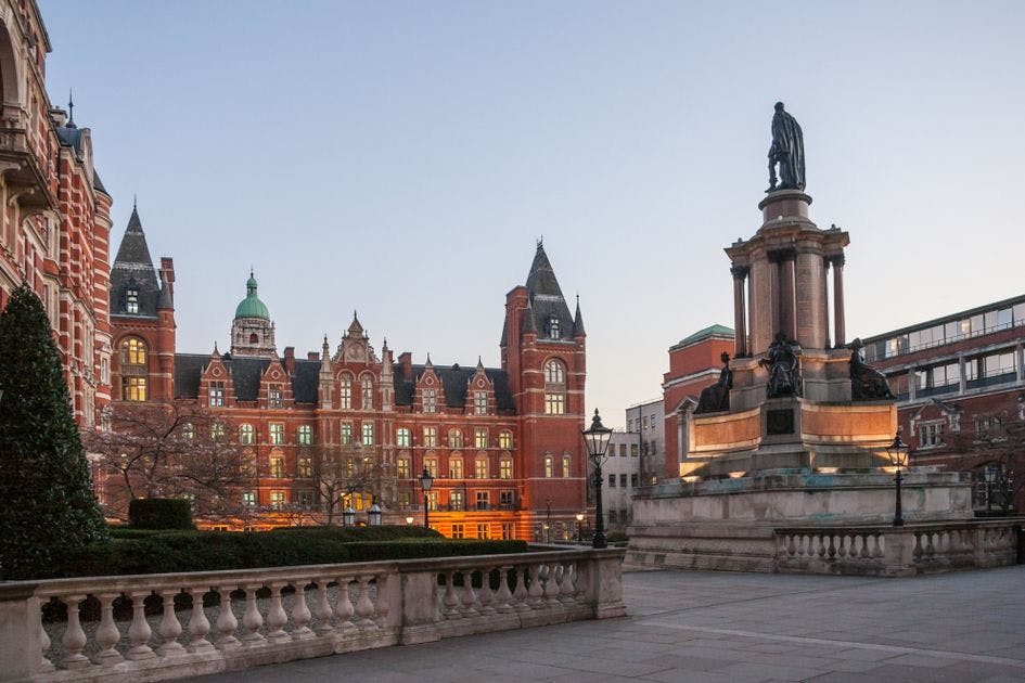 A historic building with a red brick facade and intricate architecture stands against a clear sky, accompanied by a large statue on a stone pedestal in the foreground. Best place for international student.