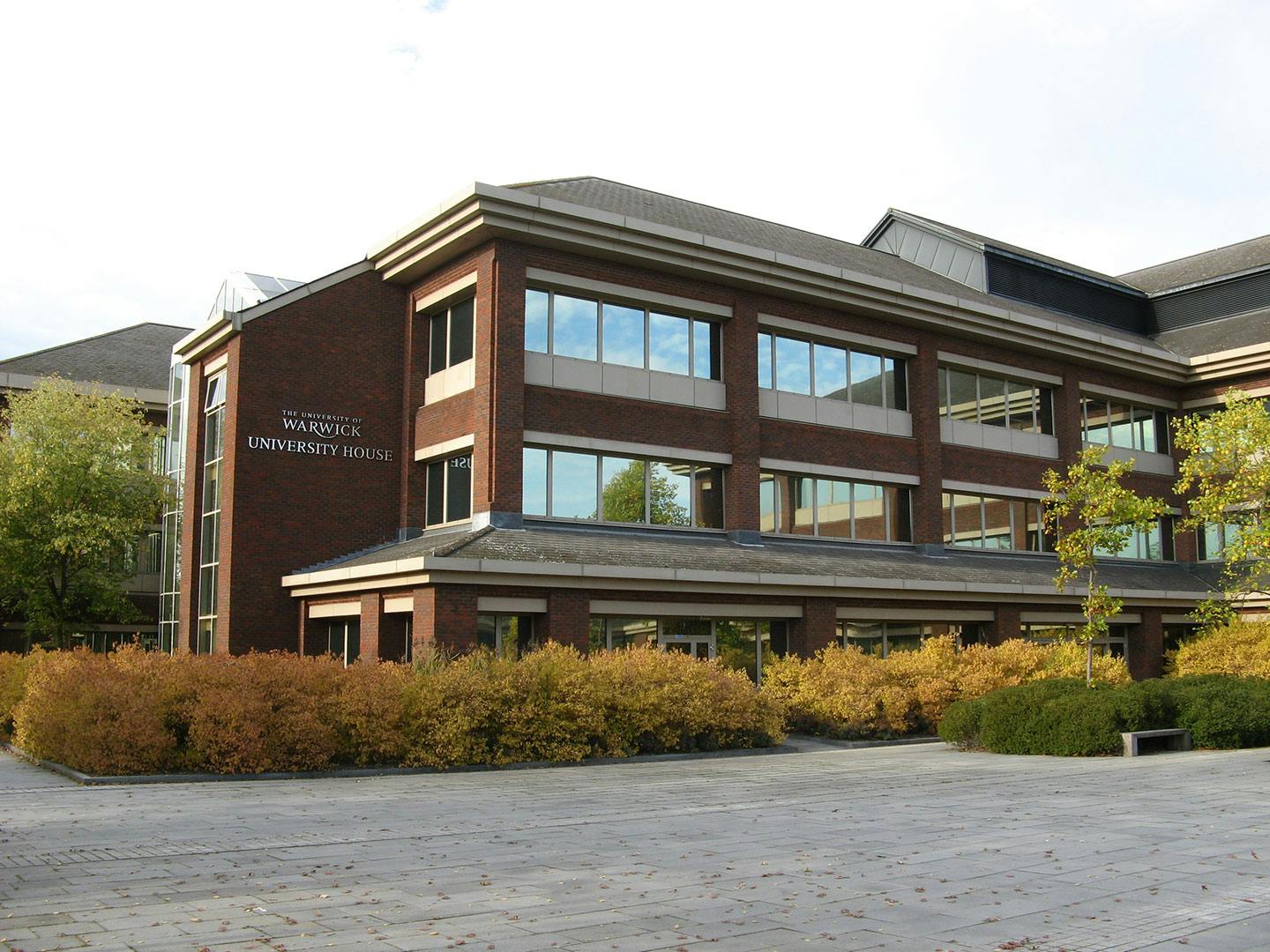 A modern brick building with large windows, identified as the University of Warwick University House, surrounded by greenery and a paved courtyard. Best place for international student. 