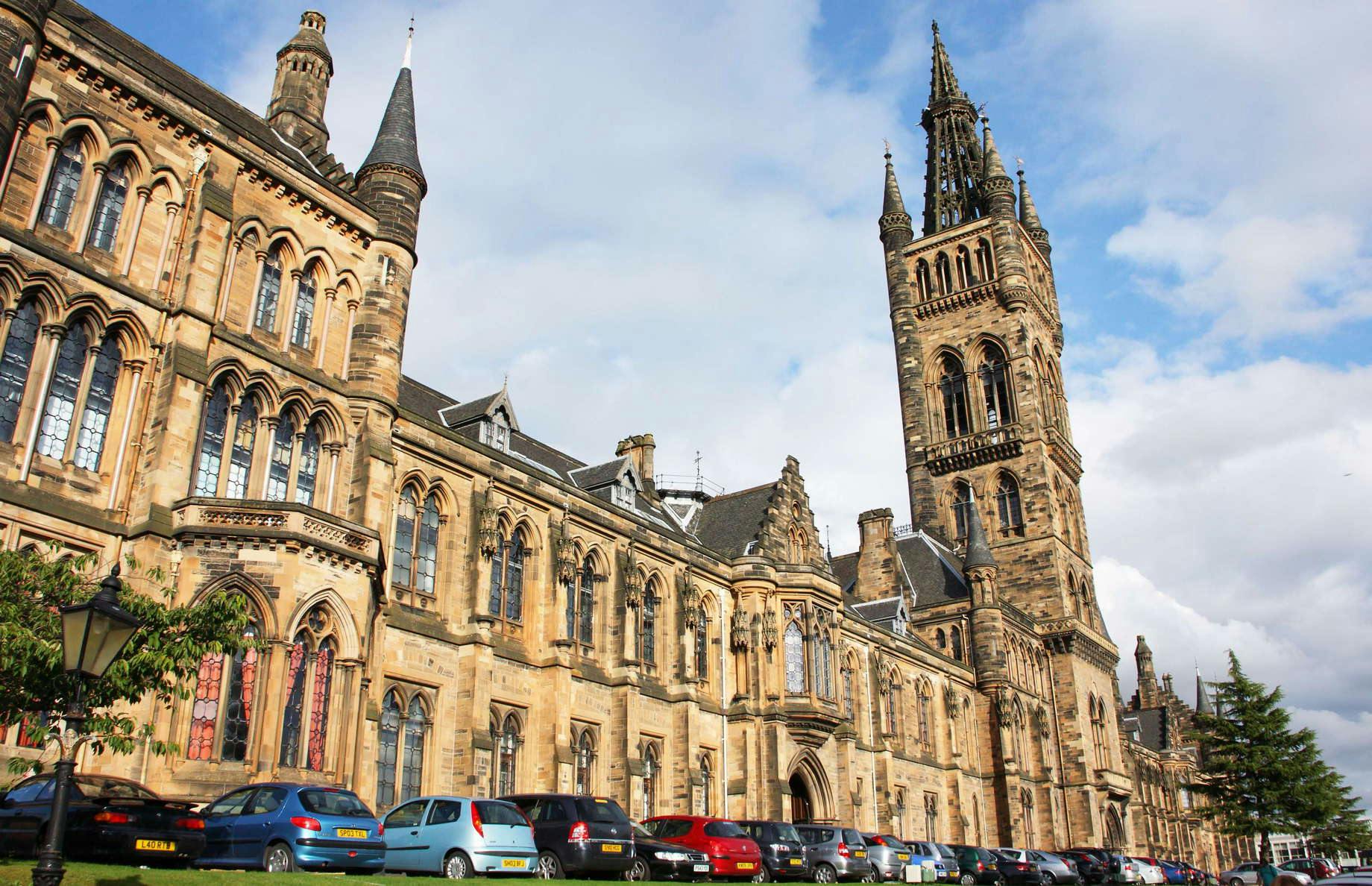 A historic stone building with ornate arches and a tall spire, with cars parked in front, set against a cloudy blue sky. Best place for international student.