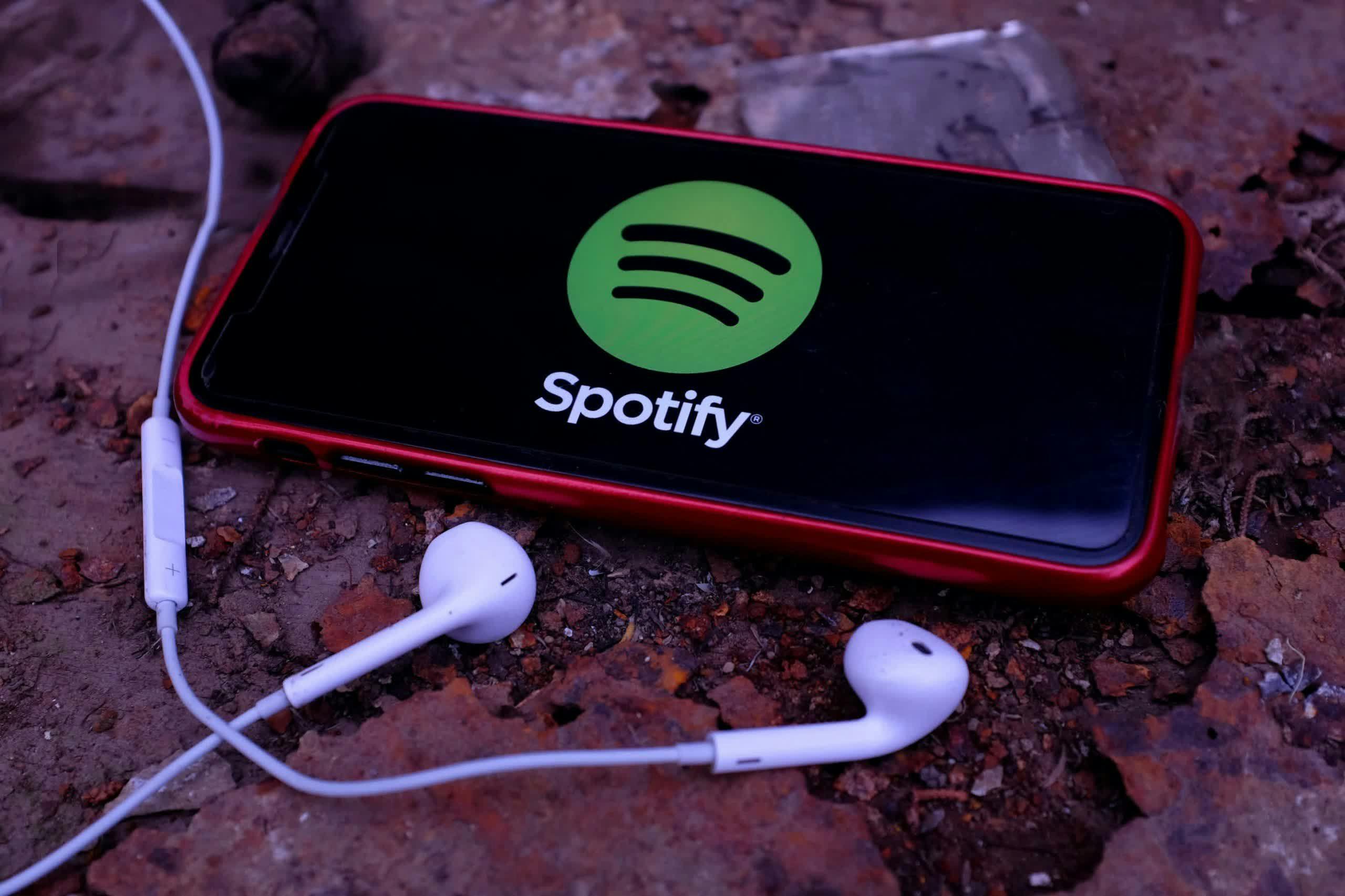 A smartphone with a red case displaying the Spotify logo on its screen lies on a textured surface, with white earbuds placed nearby.