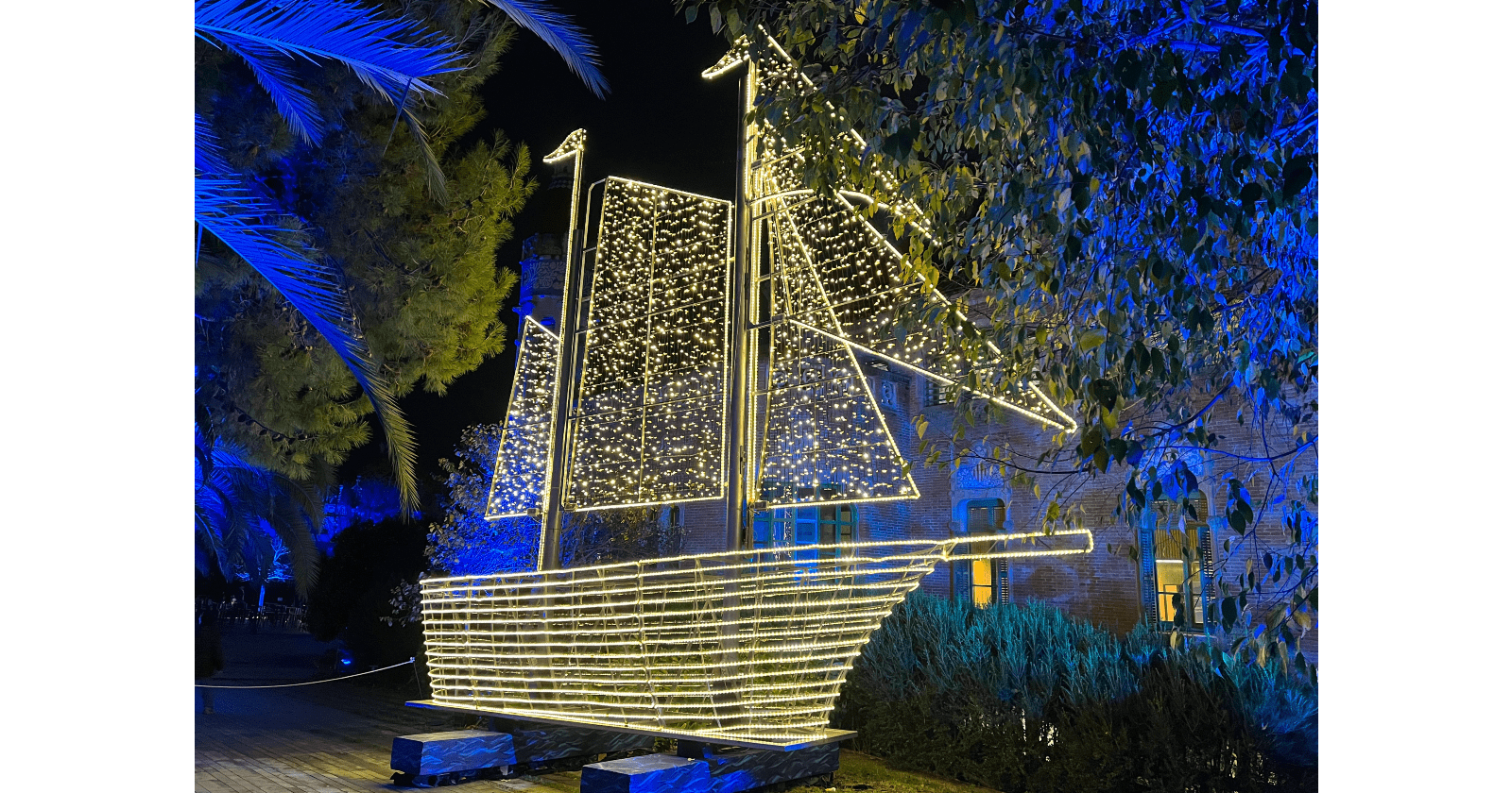 A festive ship sculpture is adorned with twinkling lights, set against a backdrop of colourful trees and a brick building at night.