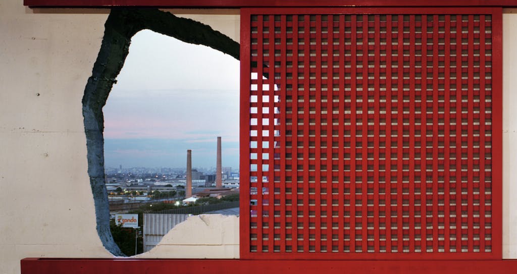 Close up of a hole on the wall of a building with a red metallic window frame around the hole; highlighting the contrast between the organic shape of the hole versus the geometric shape of the window frame
