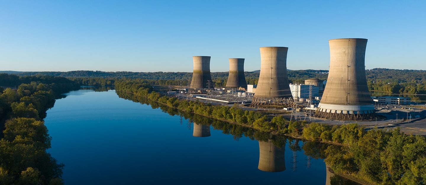 Wide photograph of the Three Mile Island site in Pennsylvania, showing cooling towers, water and wooded surroundings, published by Constellation for the Crane Clean Energy Center.